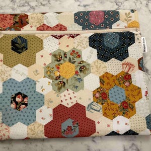 May include: A quilted fabric pouch with a zip closure. The pouch features a patchwork design of hexagonal shapes in various floral patterns and colours, including blue, red, yellow, and green. The zip is beige.