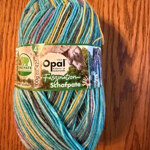 May include: A skein of variegated yarn in shades of turquoise, yellow, grey, and pink. The label features the text Opal 4-fach Pullover- & Sockenwolle, Faszination Schafpate, 100g ca. 425m, and an illustration of a sheep in a green field.