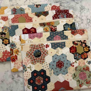 May include: Four handmade zippered pouches with a patchwork design. The pouches feature a variety of floral and geometric patterns in shades of cream, red, blue, and yellow. Each pouch has a beige zipper and a small white tag.