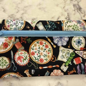 Op de afbeelding: Een rechthoekig tasje met rits op een zwarte stoffen ondergrond, voorzien van een kleurrijke print met borduurringen, garenklosjes, scharen en speldenkussens. Een lichtblauwe rits aan de bovenzijde en een wit label met Dragonfly Realm.