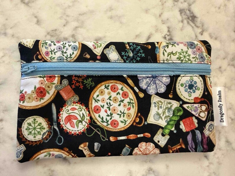 Op de afbeelding: Een rechthoekig tasje met rits op een zwarte stoffen ondergrond, voorzien van een kleurrijke print met borduurringen, garenklosjes, scharen en speldenkussens. Een lichtblauwe rits aan de bovenzijde en een wit label met Dragonfly Realm.