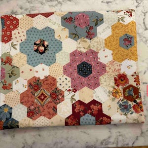 May include: A quilted fabric item with a floral pattern. The design features a patchwork of hexagonal shapes in various colours, including red, blue, yellow, and pink, with floral and geometric patterns. The item has a white label on the side.