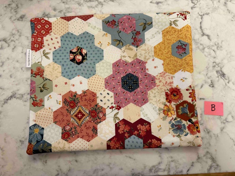 May include: A quilted fabric item with a floral pattern. The design features a patchwork of hexagonal shapes in various colours, including red, blue, yellow, and pink, with floral and geometric patterns. The item has a white label on the side.