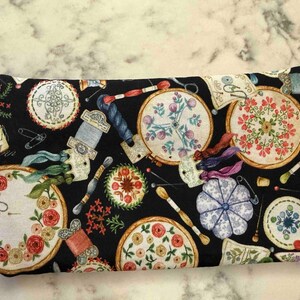 Op de afbeelding: Een rechthoekig stoffen tasje met een zwarte achtergrond en een herhalend patroon van borduurringen, kleurrijke bloemmotieven, garenklosjes, naalden en een blauw speldenkussen. Een wit stoffen label is aan de linkerzijkant bevestigd.
