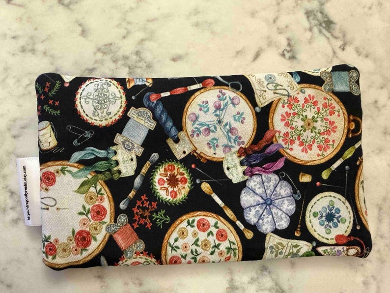 Op de afbeelding: Een rechthoekig stoffen tasje met een zwarte achtergrond en een herhalend patroon van borduurringen, kleurrijke bloemmotieven, garenklosjes, naalden en een blauw speldenkussen. Een wit stoffen label is aan de linkerzijkant bevestigd.