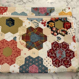 May include: A patchwork fabric pouch with a beige zip. The pouch features a design of hexagonal shapes in various floral and patterned fabrics, including red, blue, and green. The overall design is a mix of floral and geometric patterns.