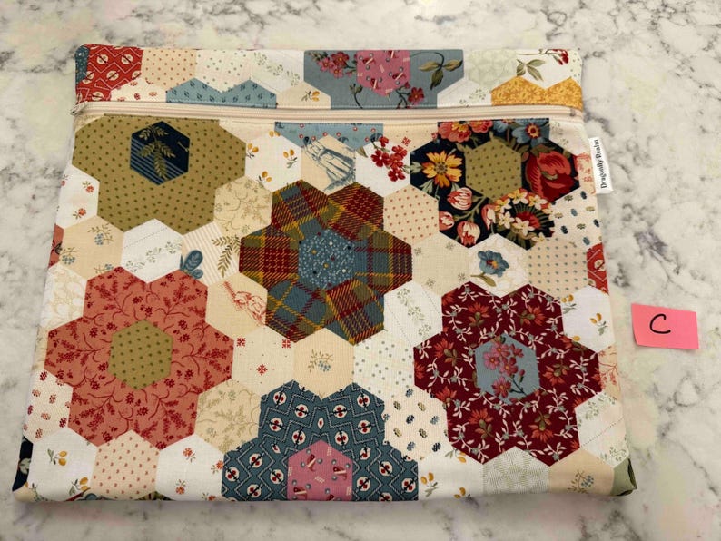 May include: A patchwork fabric pouch with a beige zip. The pouch features a design of hexagonal shapes in various floral and patterned fabrics, including red, blue, and green. The overall design is a mix of floral and geometric patterns.