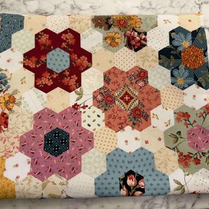 May include: A patchwork fabric item with a hexagonal design. The pattern incorporates floral and geometric motifs in red, pink, blue, and yellow hues. A label is visible on the side, and a pink tag displays the letter "C".