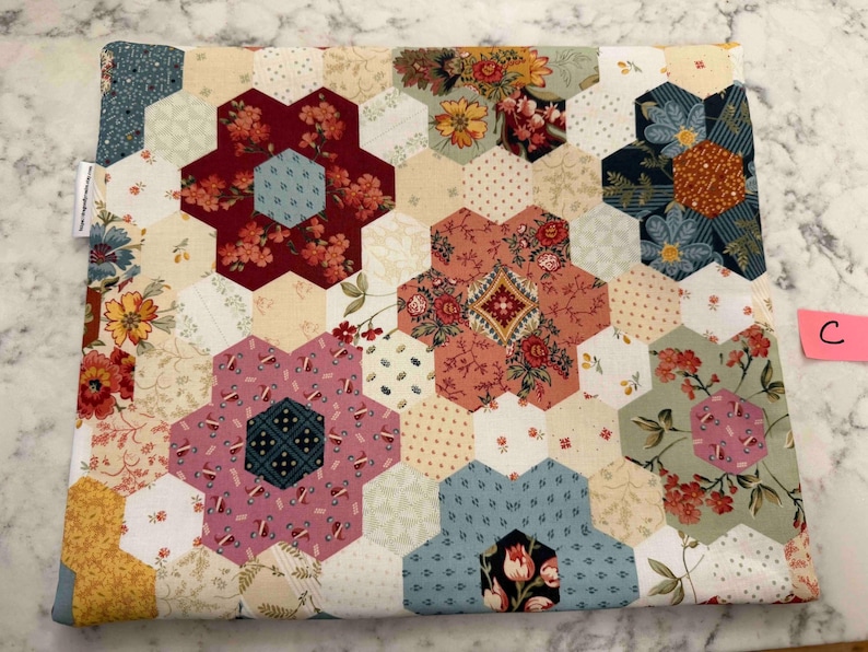 May include: A patchwork fabric item with a hexagonal design. The pattern incorporates floral and geometric motifs in red, pink, blue, and yellow hues. A label is visible on the side, and a pink tag displays the letter "C".