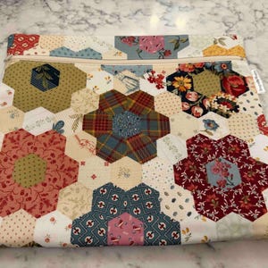 May include: A quilted pouch with a floral design. The pouch is constructed from various hexagonal fabric pieces in red, blue, green, and cream, arranged in a flower pattern. A beige zip runs across the top. A small pink tag with the letter "C" is attached.
