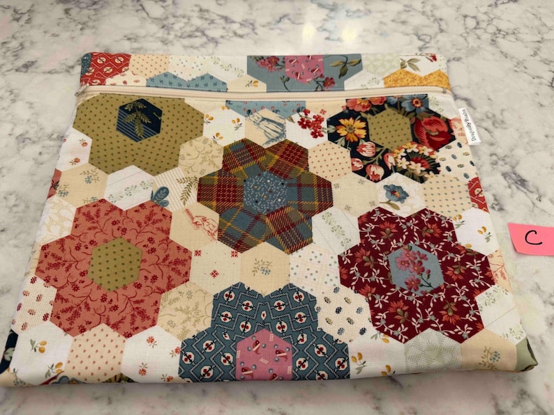 May include: A quilted pouch with a floral design. The pouch is constructed from various hexagonal fabric pieces in red, blue, green, and cream, arranged in a flower pattern. A beige zip runs across the top. A small pink tag with the letter "C" is attached.