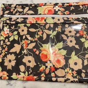 May include: A rectangular pouch with a floral pattern featuring roses and other flowers in peach, cream, and yellow against a black background. The pouch has a clear plastic window and a white zipper closure.