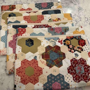 May include: Four quilted zipper pouches with a patchwork design. The pouches feature floral and geometric patterns in red, blue, green, and cream. Each pouch has a beige zip.