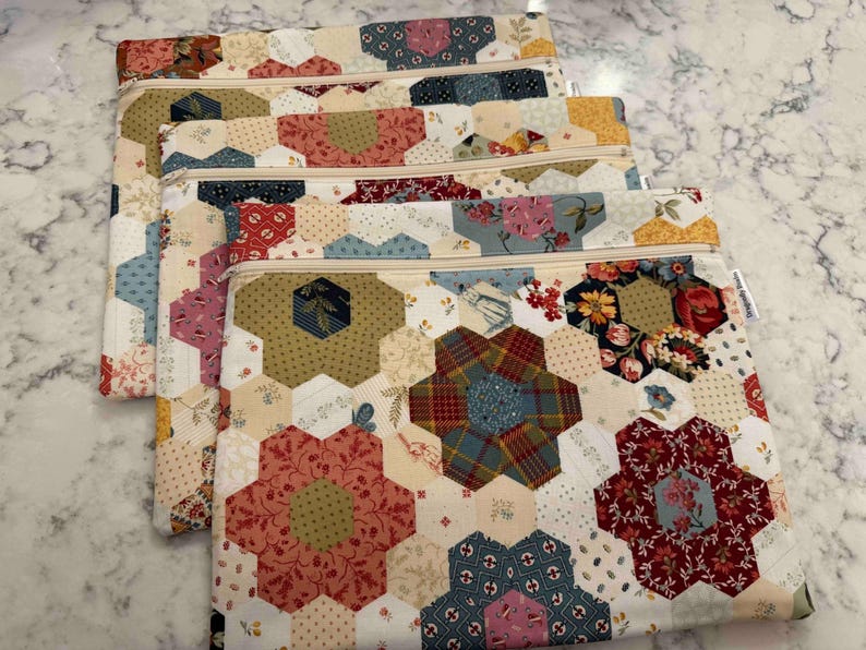 May include: Four quilted zipper pouches with a patchwork design. The pouches feature floral and geometric patterns in red, blue, green, and cream. Each pouch has a beige zip.