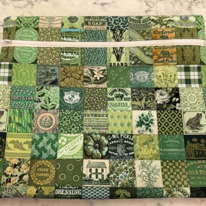 May include: A green zippered pouch with a patchwork design. The patchwork is composed of various green squares with vintage-style graphics and text, including floral and botanical motifs. The zipper is white.