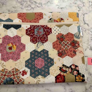 May include: A quilted fabric pouch with a floral hexagon pattern in shades of pink, red, blue, and yellow. The pouch has a beige zip and a small tag on the side. The overall design is reminiscent of a patchwork quilt.