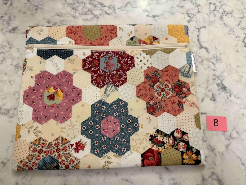 May include: A quilted fabric pouch with a floral hexagon pattern in shades of pink, red, blue, and yellow. The pouch has a beige zip and a small tag on the side. The overall design is reminiscent of a patchwork quilt.