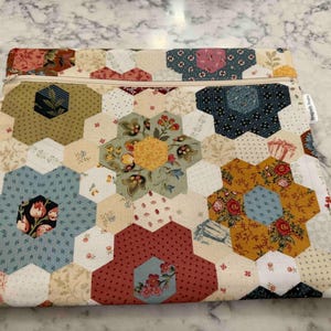 May include: A patchwork pouch with a floral hexagon design. The pouch uses fabrics in blue, red, yellow, and cream. A zip closure is along the top edge. A small pink tag with the letter "A" is visible.