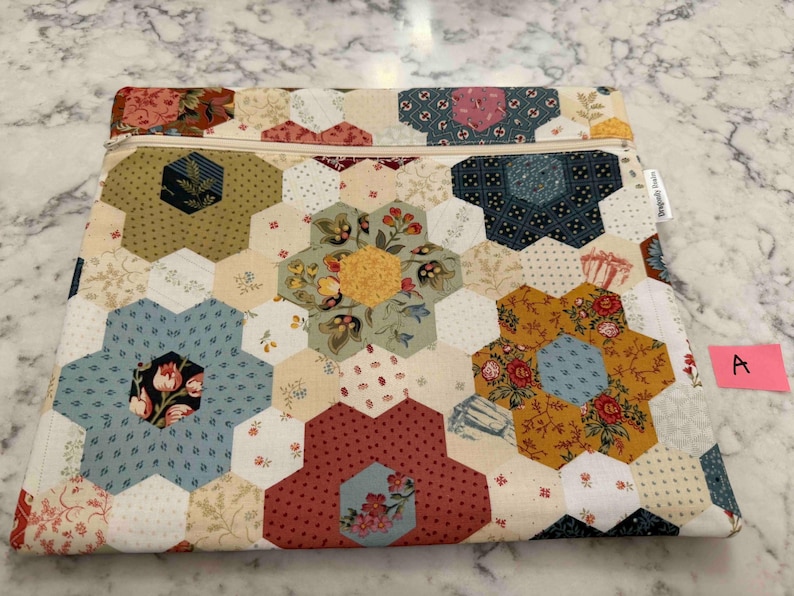 May include: A patchwork pouch with a floral hexagon design. The pouch uses fabrics in blue, red, yellow, and cream. A zip closure is along the top edge. A small pink tag with the letter "A" is visible.