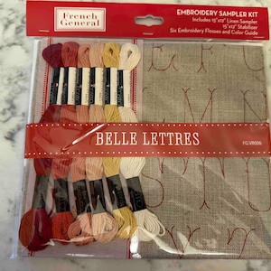 May include: A French General Belle Lettres embroidery sampler kit in clear plastic packaging. The kit includes six skeins of embroidery floss in shades of red, pink, and yellow, and a 15 by 12 inch linen sampler with red printed letters.