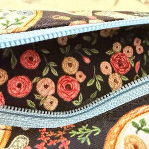 Op de afbeelding: Een stoffen etui met een donkere achtergrond en een bloemenpatroon van rode en beige rozen en groene bladeren. Het etui is voorzien van een lichtblauwe rits die langs de bovenkant loopt en contrasteert met de donkere bloemenstof.