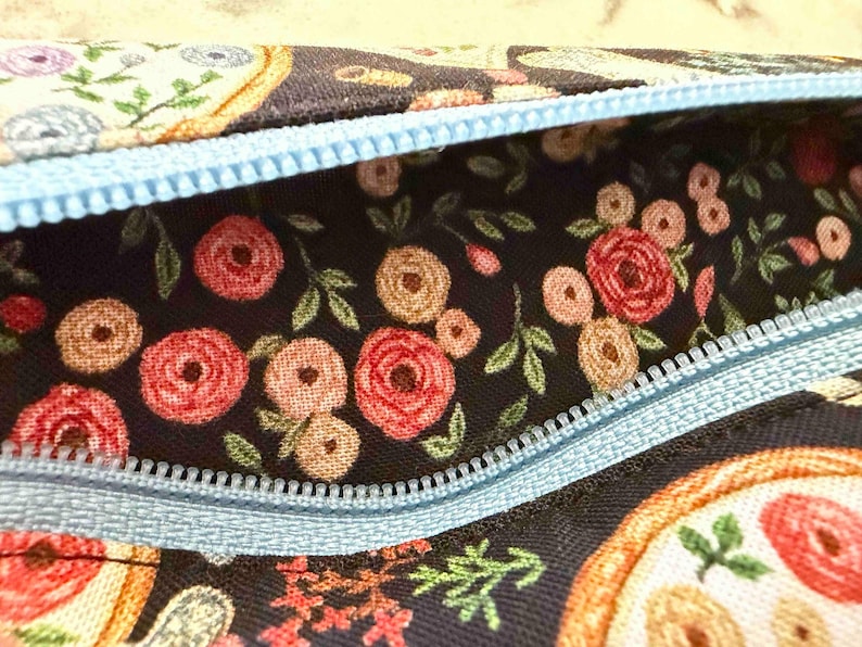 Op de afbeelding: Een stoffen etui met een donkere achtergrond en een bloemenpatroon van rode en beige rozen en groene bladeren. Het etui is voorzien van een lichtblauwe rits die langs de bovenkant loopt en contrasteert met de donkere bloemenstof.