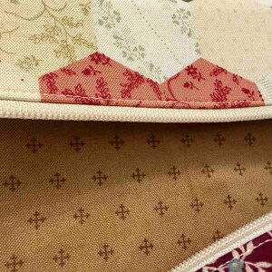 May include: Close-up of a quilted fabric with a cream-coloured zip. The fabric features a patchwork design with floral and geometric patterns in shades of red, cream, and teal. The interior lining is a tan colour with a repeating cross pattern.