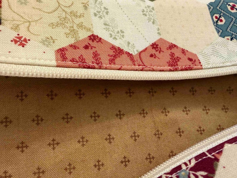 May include: Close-up of a quilted fabric with a cream-coloured zip. The fabric features a patchwork design with floral and geometric patterns in shades of red, cream, and teal. The interior lining is a tan colour with a repeating cross pattern.