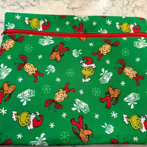 May include: A rectangular green fabric pouch featuring a repeating pattern of cartoon characters, including a green creature in a Santa hat, a blonde character with a red bow, and a brown dog, interspersed with white snowflakes and gift boxes.
