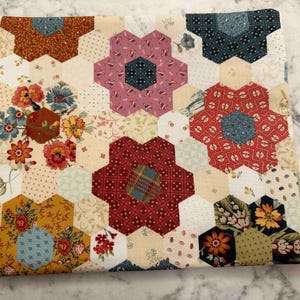 May include: A quilted fabric item with a floral pattern. The design features hexagonal shapes arranged in a patchwork style, with various floral and geometric patterns in shades of red, pink, blue, yellow, and brown. The item has a light beige background.