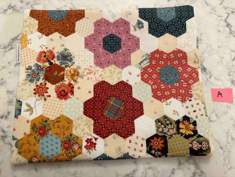 May include: A quilted fabric item with a floral pattern. The design features hexagonal shapes arranged in a patchwork style, with various floral and geometric patterns in shades of red, pink, blue, yellow, and brown. The item has a light beige background.