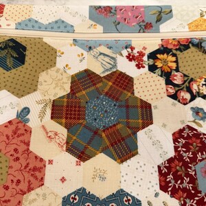 May include: A quilted pouch with a zip closure, featuring a patchwork design. The hexagonal fabric pieces display floral and patterned prints in red, blue, yellow, and cream. A handcrafted item.