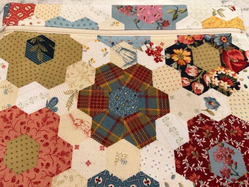 May include: A quilted pouch with a zip closure, featuring a patchwork design. The hexagonal fabric pieces display floral and patterned prints in red, blue, yellow, and cream. A handcrafted item.