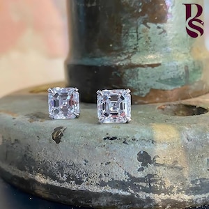 May include: A pair of square-cut diamond stud earrings. The diamonds are set in a silver or white gold setting. The earrings are displayed on a textured, metallic surface.