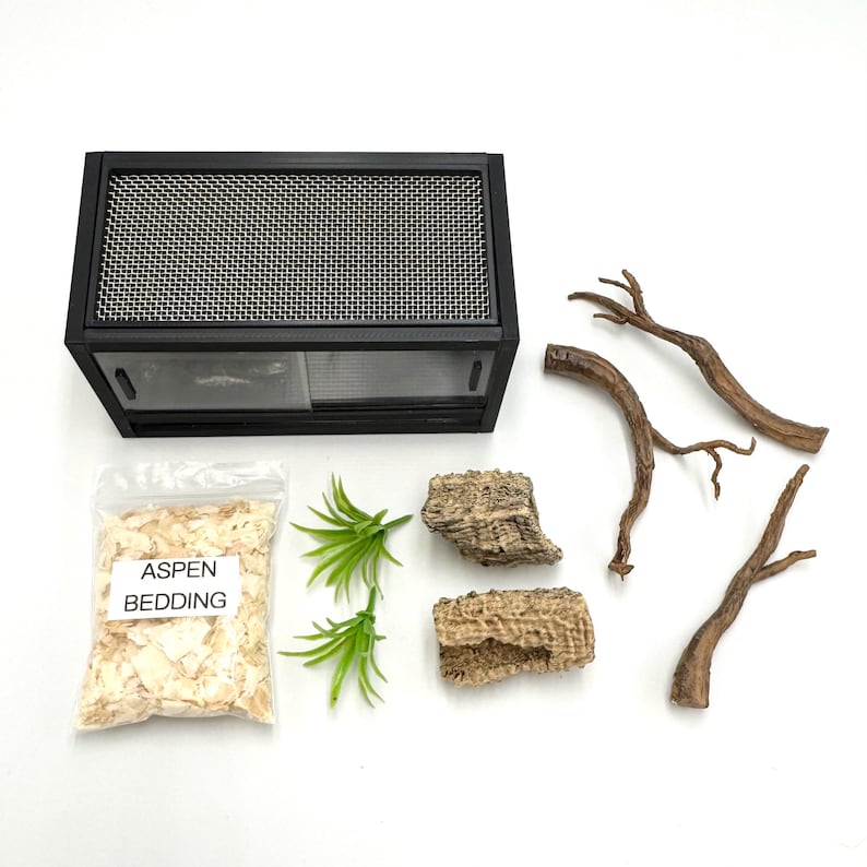 May include: A black terrarium with a mesh top, aspen bedding, decorative branches, and faux greenery. The aspen bedding is in a clear bag with the text "Aspen Bedding". The terrarium has a sliding glass door.