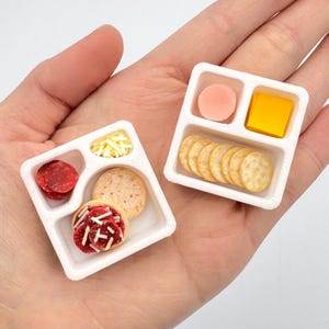 May include: Miniature charcuterie boards with various food items. One board features pepperoni, crackers, and a decorated cracker. The other board has a slice of ham, cheese, and crackers. The boards are white and held in a hand.