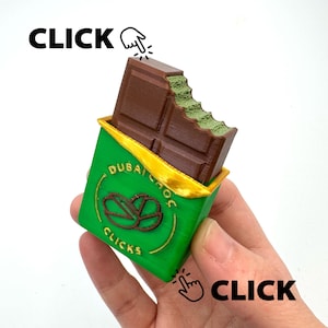 Può includere: Una barretta di cioccolato stampata in 3D con un morso, in un involucro verde con dettagli dorati. L'involucro presenta il testo "DUBAI CHOC CLICKS" e un logo. La parola "CLICK" appare in testo nero con un'icona a forma di dito.