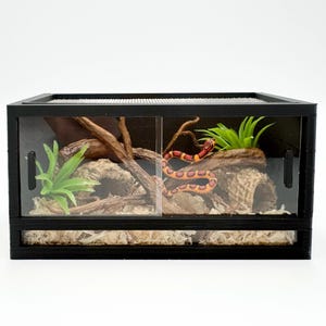 May include: A black terrarium with a mesh top and sliding glass doors. Inside, a decorative habitat features a snake, faux plants, and wood accents. The terrarium is designed for reptile or amphibian housing.