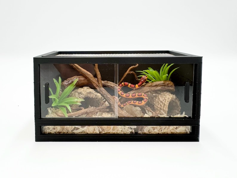 May include: A black terrarium with a mesh top and sliding glass doors. Inside, a decorative habitat features a snake, faux plants, and wood accents. The terrarium is designed for reptile or amphibian housing.