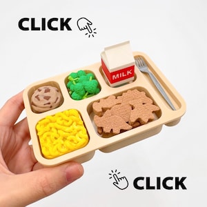 Puede incluir: Una bandeja de almuerzo beige en miniatura con compartimentos llenos de una galleta con chispas de chocolate, brócoli, un cartón de leche, galletas con forma de dinosaurio y fideos amarillos. Un tenedor pequeño descansa en la bandeja. La palabra "CLICK" aparece en texto negro.