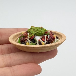May include: Miniature wooden bowl filled with a colorful salad. The salad includes black beans, corn, red tomatoes, green peppers, and a dollop of white cream, topped with a vibrant green guacamole. The bowl is held in a hand.