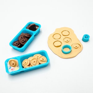 May include: A craft set with miniature donuts and a dough sheet. The set includes a blue container with chocolate donuts, a blue container with frosted donuts, and a sheet of dough with donut cutouts. A blue donut cutter is also included.