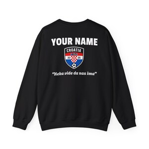Może przedstawiać: Czarna bluza z białym napisem "YOUR NAME" nad herbem Chorwacji. Herb zawiera czerwono-biały wz&oacute;r w szachownicę, piłkę nożną i napis "20 25". Poniżej napis "Neka vide da nas ima".
