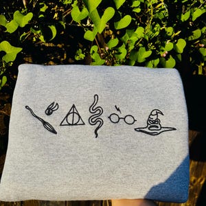 May include: A gray sweatshirt featuring black embroidered symbols from a popular fantasy series. The symbols include a broom, lightning bolt, triangle, snake, glasses, and a sorting hat. The sweatshirt is displayed against a backdrop of green foliage.