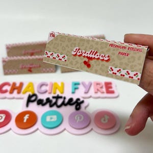 May include: A pack of Jordiloos premium rolling paper is held in front of a pink and white sign that says "Chacin Fyre Parties". The rolling paper has a tan leopard print design with cherry accents and the words "Jordiloos" and "Premium Rolling Paper".