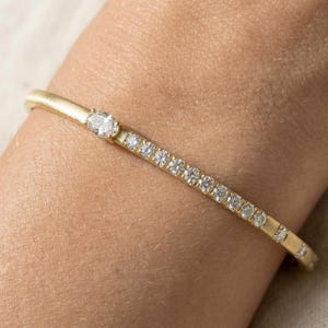 Puede incluir: Un brazalete de oro con una fila de pequeños diamantes brillantes. El brazalete tiene un diseño simple y elegante con un diamante más grande en la parte superior. El brazalete se lleva en la muñeca.