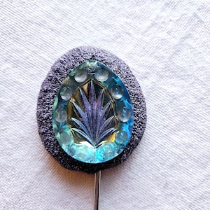 Peut inclure: Épingle à chapeau vintage avec un bijou en verre bleu et vert irisé en forme de goutte d'eau. Le bijou présente un motif floral sculpté et est monté sur un support texturé gris foncé. La broche a une tige en métal argenté.