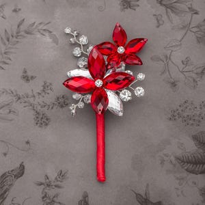 May include: A floral boutonniere featuring two large red crystal flowers with marquise-cut petals, accented by smaller clear crystal sprays and silver wire branches. The stem is wrapped in a smooth red satin ribbon, creating a structured design.