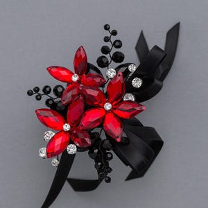 Peut inclure: Une broche florale composée de trois fleurs en cristal rouge vif aux pétales taillés en marquise. L'arrangement est agrémenté de petits strass transparents, de brins de perles noires et d'un nœud en ruban de satin noir.