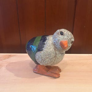 May include: A ceramic duck figurine with a speckled gray body, a black and green striped back, and orange feet and beak. The duck is posed in a standing position, with its head turned slightly to the side. The figurine is on a wooden surface.